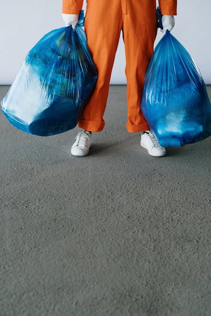 A person standing on a grey indoor or outdoor surface, holding two large blue transparent rubbish bags, one in each hand. The individual is wearing orange overalls or work trousers, white sneakers, and grey gloves. The bags are filled with waste materials, and their contents are partially visible through the plastic, appearing to contain household or general waste. The background is plain and neutral, with no identifiable environment, emphasizing the act of rubbish collection which may relate to private waste disposal or alternative rubbish removal services such as those offered by Rubbish Removal Ilford. The scene depicts a typical scene of waste handling that could be associated with independent clearance, highlighting the importance of professional rubbish removal services for efficient waste management.
