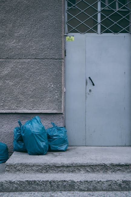 In front of a grey concrete building with a textured surface, several blue rubbish bags made of plastic are placed on a stepped concrete pavement. The bags appear full and are gathered together near the corner of the building, close to a grey metal door with a black handle, which is secured with a lever latch. Above the door, there is a section of glass blocks with a diamond pattern allowing natural light into the interior space, partially visible in the background. A small yellow warning or identification label is affixed to the corner of the building near the door frame. The scene is outdoors, with the grey tones of the building and pavement contrasting against the vibrant blue rubbish bags, reflecting a typical situation where private waste collection or on-site clearance by rubbish removal services might be involved. This setup is representative of waste temporarily stored prior to collection, fitting within a context of non-standard, independent rubbish disposal outside of municipal services.