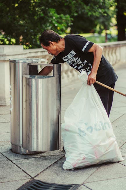 A person standing on a grey indoor or outdoor surface, holding two large blue transparent rubbish bags, one in each hand. The individual is wearing orange overalls or work trousers, white sneakers, and grey gloves. The bags are filled with waste materials, and their contents are partially visible through the plastic, appearing to contain household or general waste. The background is plain and neutral, with no identifiable environment, emphasizing the act of rubbish collection which may relate to private waste disposal or alternative rubbish removal services such as those offered by Rubbish Removal Ilford. The scene depicts a typical scene of waste handling that could be associated with independent clearance, highlighting the importance of professional rubbish removal services for efficient waste management.