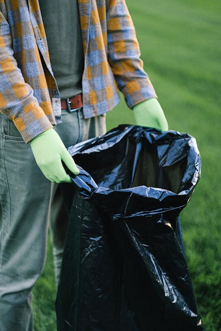 A person wearing a plaid shirt in shades of orange, blue, and gray, along with a green jacket and light gray trousers, is standing outdoors on a grassy area. They are holding open a large black rubbish bag made of thick plastic with both hands; the bag appears to be partially filled and ready for waste disposal. The individual is wearing light green gloves, suggesting they are engaging in rubbish collection or waste management. The background consists of a well-maintained grassy lawn, indicative of a garden, park, or open outdoor space. The natural lighting highlights the contrasting colors of the clothing and the bright green grass, emphasizing an environment linked to outdoor waste collection or rubbish removal activities, as carried out by local services such as Rubbish Removal Ilford. This scene demonstrates a typical scene of private waste handling activity that may relate to alternative waste disposal efforts outside of municipal rubbish collection services, emphasizing the importance of proper rubbish disposal and responsible waste management practices.