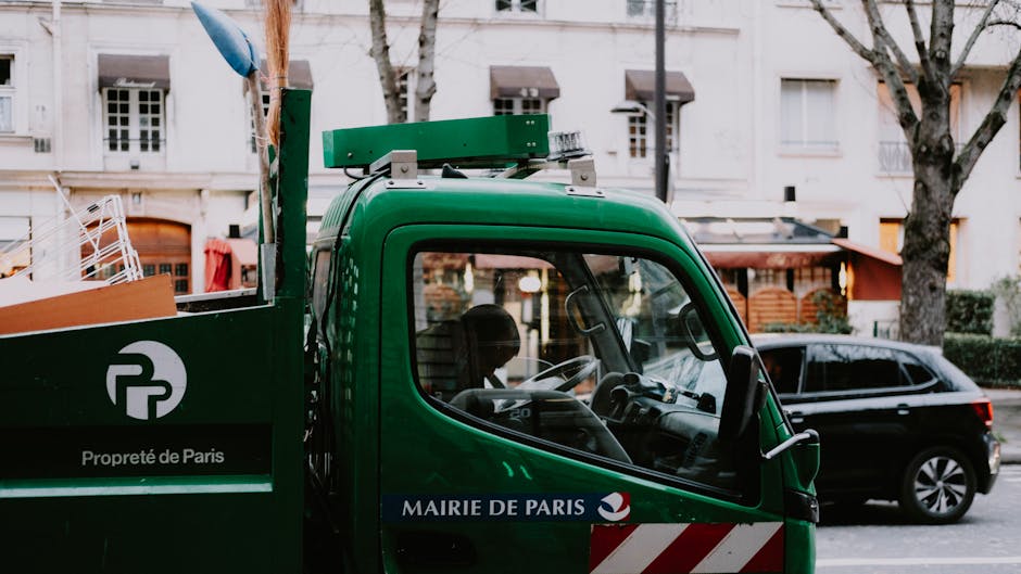 A green waste collection truck belonging to Mairie de Paris, with branding indicating it is part of Paris city rubbish services, is parked on a street with a residential area in the background. The truck's side panel displays a logo with the letter 'P' and the text 'Propreté de Paris', along with a black and white stripe pattern near the lower section. The truck has a compact, boxy cab with a large, curved windshield and side window, providing visibility into the driver's seat area. The environment includes a black car parked behind the truck and leafy trees with bare branches, suggesting late autumn or winter, as well as a row of multi-story white buildings with small windows and decorative architectural details. The overall scene appears to be during daylight hours with natural light illuminating the outer urban setting, and the truck is positioned close to a sidewalk, ready for independent waste collection or on-site rubbish removal, supporting services offered by rubbishremovalilford.co.uk in the context of alternative waste handling methods.