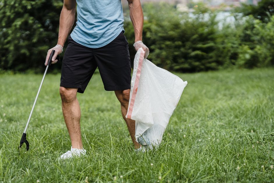 A person standing outdoors in a grassy area, holding a white trash bag with a pink stripe along the top edge in their right hand, while their left hand holds a litter picker tool with a black gripping end. The individual is dressed in a light blue T-shirt, black shorts, and white sneakers, and is wearing light-colored gloves. The background features blurred green foliage and trees, suggesting a park or garden environment. This scene illustrates private waste collection or cleanup efforts, possibly related to alternative rubbish removal services, with the individual engaged in collecting litter for disposal or recycling. The lighting is natural, and the scene appears calm, emphasizing a responsible approach to waste management and outdoor maintenance.