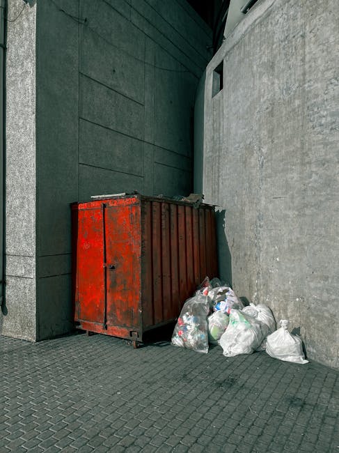 A large, red industrial waste skip with visible rust and peeling paint is positioned against a grey concrete wall in an outdoor urban setting, adjacent to a building with modern, dark green cladding. Surrounding the base of the skip are multiple white and clear plastic garbage bags, some partially torn open, revealing assorted household waste such as plastic bottles and packaging. The ground is paved with dark grey, interlocking bricks, and a narrow alleyway or corner is formed by the building and the wall, with a small, rectangular window visible high up on the wall. The overall scene depicts a typical waste collection point where private or independent rubbish removal services, like those offered by Rubbish Removal Ilford, might handle such refuse, illustrating a practical example of on-site rubbish disposal rather than public curbside collection.