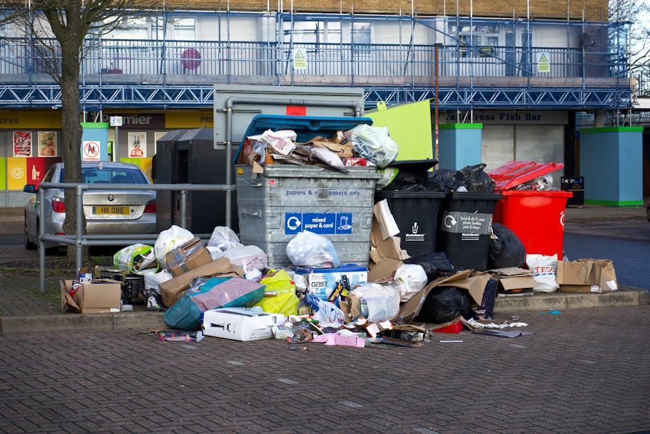 A large, red industrial waste skip with visible rust and peeling paint is positioned against a grey concrete wall in an outdoor urban setting, adjacent to a building with modern, dark green cladding. Surrounding the base of the skip are multiple white and clear plastic garbage bags, some partially torn open, revealing assorted household waste such as plastic bottles and packaging. The ground is paved with dark grey, interlocking bricks, and a narrow alleyway or corner is formed by the building and the wall, with a small, rectangular window visible high up on the wall. The overall scene depicts a typical waste collection point where private or independent rubbish removal services, like those offered by Rubbish Removal Ilford, might handle such refuse, illustrating a practical example of on-site rubbish disposal rather than public curbside collection.