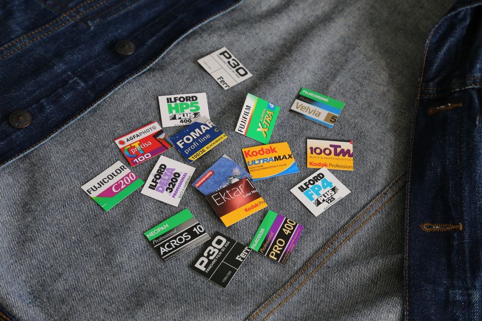 A collection of photographic film labels and packaging stickers scattered across a pair of blue denim jeans with visible orange stitch detail. The labels include brands such as Ilford, Kodak, Fujifilm, and AGFA, in various rectangular shapes with bright colors like green, red, purple, black, and white, each displaying specific film type, ISO speed, or product descriptions. The jeans are laid flat, with the labels positioned on the front thigh area, and the background appears to be a neutral surface. The scene exemplifies the sort of items that might accumulate during private or on-site disposal, relevant to rubbish removal services, with the labels demonstrating discarded photographic film packaging materials often handled by independent waste collection providers like Rubbish Removal Ilford in contexts such as garden waste removal or general rubbish clearance. Lighting is even, highlighting the texture of the denim and the glossy finish of the labels, supporting an accurate, professional, and neutral description of the physical objects visually present.