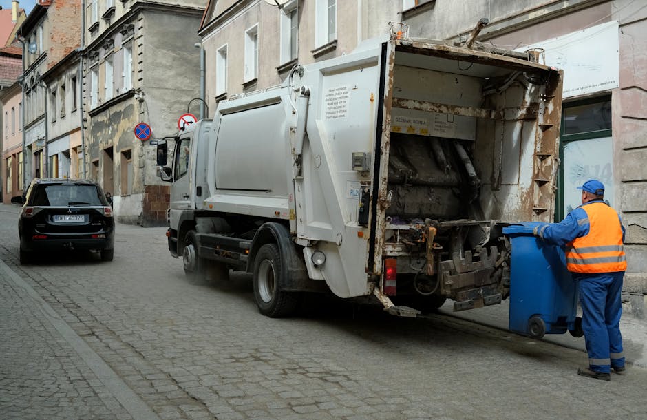A white waste collection truck with an open rear hopper, showing a rusted metal interior and mechanical components, is parked on a narrow cobblestone street alongside nearby residential and commercial buildings. A worker dressed in a blue uniform and high-visibility orange vest, wearing a blue cap, is seen pushing a blue wheelie bin toward the truck's rear to unload waste. The scene is outdoors during daytime with muted natural lighting, and additional vehicles, including a black car parked further along the street, are visible in the background. The setting suggests a private waste collection or rubbish removal service operating in an urban area, consistent with alternative waste handling options such as on-site rubbish collection by services like Rubbish Removal Ilford, situated on high roads and residential streets for flats and townhouses.