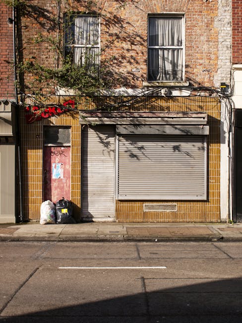 The image depicts the front facade of a mixed-use building with a brick upper section, featuring two vertically aligned windows with white frames and partially drawn curtains. The ground floor consists of a small commercial space with a roll-up metal shutter in a closed position, painted in a light grey tone, centered beneath the brickwork. To the left of the shutter, there is a pinkish-red wooden door with visible graffiti and some peeling paint, adjacent to a small area of yellow tiles that line the lower portion of the building’s exterior. On the sidewalk directly in front, three black plastic garbage bags and a white bag are placed near the wall, indicating waste disposal. Above, cable conduits and wiring are visible, running horizontally across the building’s facade. The scene is illuminated by natural daylight, casting distinct shadows of nearby tree branches onto the brick wall, suggesting an outdoor urban environment. The image relates to private waste management practices, such as alternative rubbish collection and on-site clearance, which Rubbish Removal Ilford may facilitate for local properties.