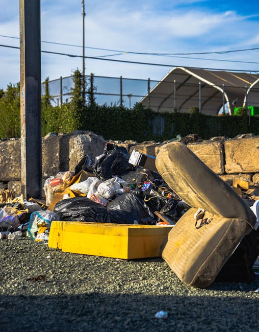 A large pile of mixed waste materials sits on a gravel surface outdoors, including black plastic garbage bags, an old, worn beige car tyre, and a yellow plastic container or tray. Behind the waste heap, a low stone wall made of roughly hewn stones runs horizontally across the image, and above it, a row of trees and a tall utility pole with overhead power lines are visible. In the background, there is a large metal-framed structure with a curved, semi-transparent roof, partially obscured by a dark mesh fence and greenery, suggesting an industrial or outdoor storage area. The scene is illuminated by natural daylight under a blue sky with some wispy clouds, capturing a snapshot of roadside rubbish that may be subject to independent removal service, consistent with private waste handling. The overall environment appears to be a designated or informal dumping site, reflecting the kind of waste typically handled by companies like Rubbish Removal Ilford, suited for on-site clearance or alternative waste collection.
