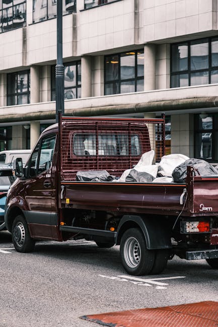 A burgundy flatbed truck parked on an urban street in front of a modern office building with large glass windows and vertical concrete panels. The truck's open cargo area is filled with various types of waste materials, including several black and white plastic bags, some appearing partially torn or loosely tied, and miscellaneous debris. The bed has a metal side railing and a grid-style protection behind the cab, both painted matching the vehicle's colour, and the rear wheel is visible beneath the load. The scene is illuminated by daylight with no visible signage or markings indicating a commercial service. The presence of the truck and waste cargo suggests it is involved in private waste collection or rubbish removal services, consistent with the offerings of companies like Rubbish Removal Ilford, providing alternative waste handling options in the area.