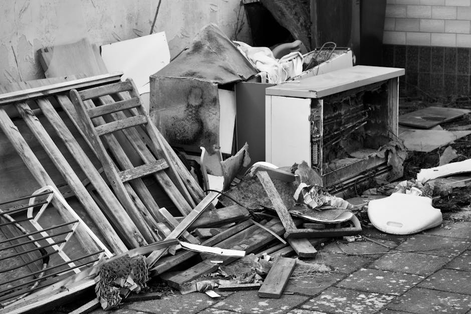 A pile of discarded household items and debris is accumulated on a paved outdoor surface near a building wall. The pile includes a wooden garden chair with slatted back and seat, leaning against other materials; a small, white, broken appliance or container partially buried among the rubbish; a damaged piece of furniture or large panel with visible wear and dirt; and various broken wooden planks, pieces of cardboard, and torn fabric. There is also a large, dark, partially covered object that appears to be a piece of furniture or appliance, with its surface stained and weathered. The debris is haphazardly arranged, with some items partially obscured behind others, and the surrounding environment shows a brick wall and a concrete or tiled surface, indicating an outdoor setting close to a building. This scene exemplifies private waste or bulky rubbish that might be collected independently from a property, highlighting the need for professional rubbish removal services like those offered by Rubbish Removal Ilford to handle such on-site clearance efficiently.