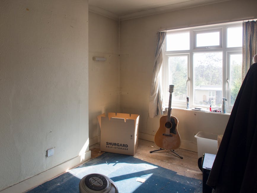 A small, empty room with beige painted walls and a wooden floor partially covered by a dark blue carpet. Near a large, white-framed double-glazed window that allows natural daylight to illuminate the space, a light blue curtain is pulled to one side. In the corner, there is a cardboard box labeled 'SHURGARD SELF-STORAGE' that appears to be used for storage or moving purposes. An acoustic guitar with a natural wood finish and six strings is propped upright on a black stand close to the window. On the windowsill, various small objects including a candle and a bottle can be seen. To the right, a white plastic storage container is placed on the floor, partially obscured by a dark coat hanging on a nearby hook or stand. A small, round robotic vacuum cleaner is situated on the floor in the foreground, with light casting soft shadows across the room. The space appears to be in the process of clearing or preparing for a rubbish removal service, with a plain environment and no furniture or furnishings present.