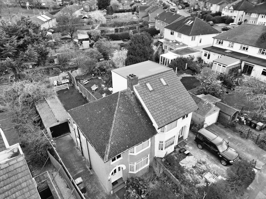 Aerial black-and-white photograph of a residential property featuring a two-storey house with a tiled pitched roof, dormer windows, and surrounding garden area. The front garden includes a driveway with an SUV parked, and a small paved pathway leading to the front entrance. Adjacent to the house, there is a detached garage and a backyard enclosed by fencing, containing a shed, garden furniture, and trees with bare branches, suggesting a season outside of summer. Several nearby houses with similar architectural styles are visible, each with varied roof types and garden arrangements. The neighborhood appears densely populated with closely spaced houses, some with visible solar panels on rooftops. The overall scene conveys a quiet suburban area with well-maintained properties, typical of private on-site waste disposal or rubbish collection services occasionally used for house clearance projects by companies like Rubbish Removal Ilford, as evidenced by the surrounding environment and structure of the properties.