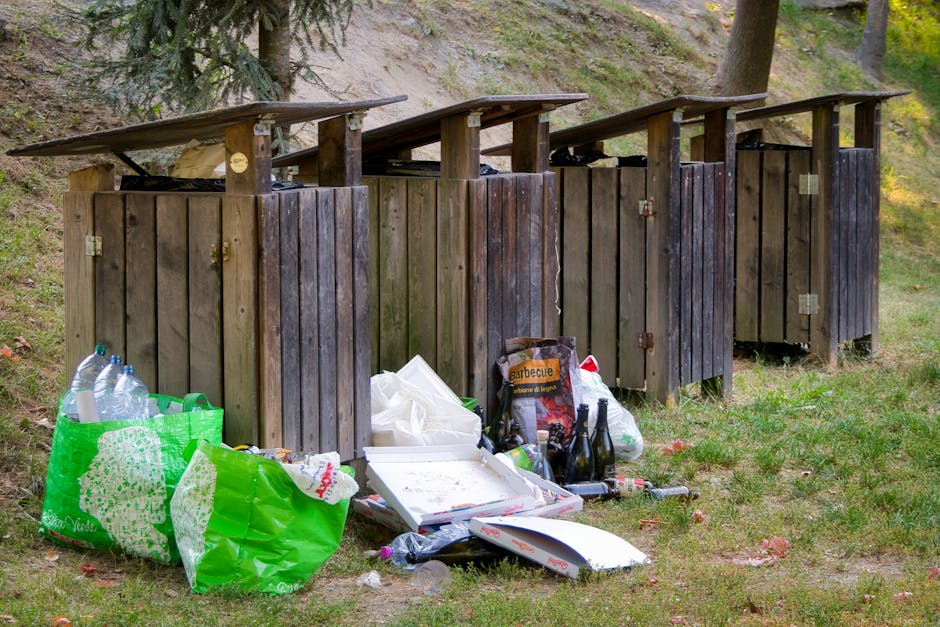 A row of four wooden outdoor waste bins with hinged flat roofs, positioned on a grassy area in front of a slight hillside with trees and foliage. The bins are weathered, dark-stained, and made of vertical wooden planks, each secured with metal hinges and locks. The leftmost bin has a green plastic bag filled with empty plastic bottles, some with transparent labels visible. In front of the bins, there are various discarded items including a white pizza box, plastic bottles, glass bottles, a black bag, and some miscellaneous waste, all loosely arranged on the grass. The background features a sloping hillside with patches of grass, small plants, and trees, casting natural daylight across the scene. The overall environment appears to be an outdoor area used for waste disposal, with evidence of littering indicative of non-official rubbish collection, aligning with services such as those offered by Rubbish Removal Ilford for private waste handling and on-site clearance.