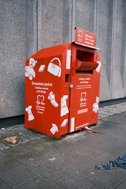 A red donation drop-off box positioned on a paved sidewalk against a grey concrete wall. The box features white illustrations of various items including a teddy bear, a handbag, a pair of shoes, clothing, and books, indicating it is for donating clothes, books, and other belongings. There is a small opening with a flap at the top for inserting items, and a separate slot below for collection. The box has printed text referencing it as a donation point for the British Heart Foundation and indicates it accepts clothing, books, and other things. The surrounding environment appears to be outdoors, with a small patch of litter or debris near the base and some blue fabric or plastic on the ground nearby. The overall setting suggests an accessible location for community donations, supporting alternative waste handling and charitable collection services like those offered by Rubbish Removal Ilford.