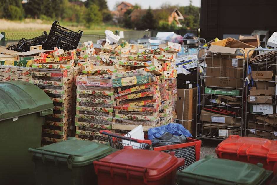 A large collection of discarded fruit and vegetable boxes, predominantly made of cardboard with bright printed labels and images of fresh produce, are stacked in an outdoor area, possibly a driveway or waste disposal site. Surrounding the boxes are various waste containers including green and red wheelie bins made of plastic with domed lids, along with metal and plastic crates filled with miscellaneous packaging materials and smaller cardboard boxes. In the background, a black metal trolley or cart, fitted with multiple shelves, holds additional cardboard boxes and packaging supplies, some still sealed with labels visible. The scene is outdoors, with natural daylight illuminating the area, and distant trees and residential buildings can be seen beyond the waste collection setup, indicating an area associated with waste management or collection managed by Rubbish Removal Ilford. The scene offers a clear view of different types of waste storage and disposal, aligning with practices of private waste handling and alternative rubbish collection methods, such as on-site clearance or independent collection, suitable for those seeking non-local authority disposal options in the Ilford area.