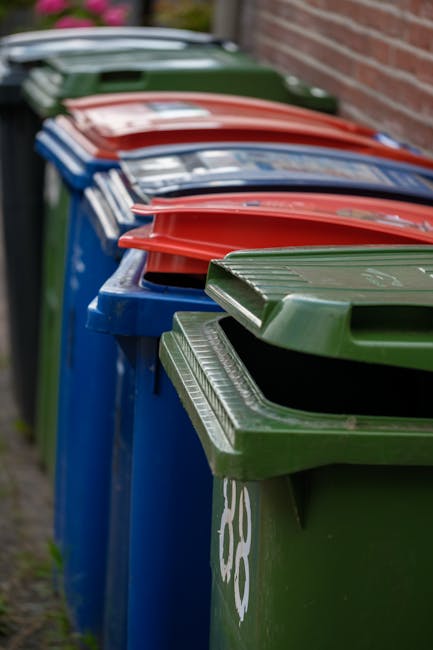A row of large residential wheelie bins is aligned along a pavement, positioned next to a brick wall. The bins are made of durable plastic with textured surfaces and have hinged lids. The closest bin in the foreground is green, with visible markings, while further down the row, there are blue, red, and dark green bins, each with different coloured lids corresponding to waste categories. The lids are closed, and the bins are standing upright, tightly packed side-by-side. The background features a blurred view of additional bins and greenery, suggesting an outdoor location typical for rubbish collection points. The lighting appears natural and overcast, providing even illumination without shadows, consistent with weekday rubbish disposal routines in residential areas. This scene exemplifies standard arrangements for private waste disposal, which waste management services like Rubbish Removal Ilford facilitate through flexibly handling alternative waste collection outside of council-authorized curbside pickups, ensuring proper segregation and disposal of various waste types.