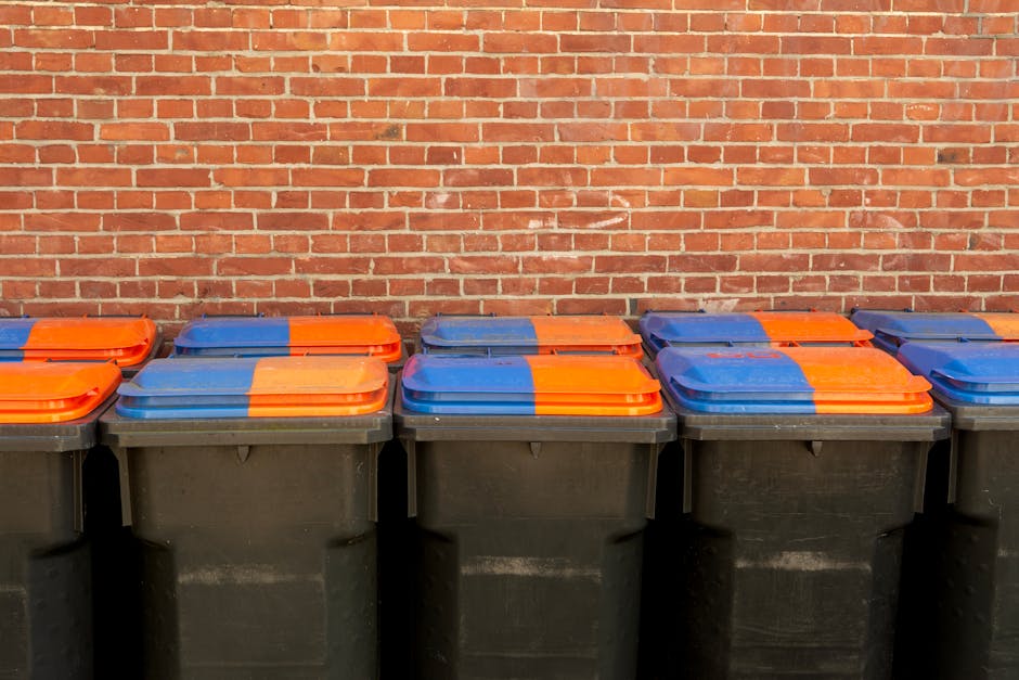 A large collection of discarded fruit and vegetable boxes, predominantly made of cardboard with bright printed labels and images of fresh produce, are stacked in an outdoor area, possibly a driveway or waste disposal site. Surrounding the boxes are various waste containers including green and red wheelie bins made of plastic with domed lids, along with metal and plastic crates filled with miscellaneous packaging materials and smaller cardboard boxes. In the background, a black metal trolley or cart, fitted with multiple shelves, holds additional cardboard boxes and packaging supplies, some still sealed with labels visible. The scene is outdoors, with natural daylight illuminating the area, and distant trees and residential buildings can be seen beyond the waste collection setup, indicating an area associated with waste management or collection managed by Rubbish Removal Ilford. The scene offers a clear view of different types of waste storage and disposal, aligning with practices of private waste handling and alternative rubbish collection methods, such as on-site clearance or independent collection, suitable for those seeking non-local authority disposal options in the Ilford area.
